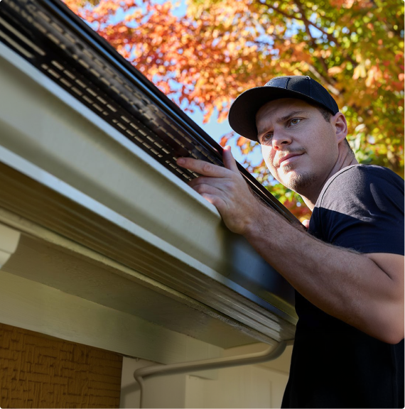 A man wearing a black baseball cap and t-shirt installs a perforated metal gutter guard on a residential home, with bright autumn foliage visible in the background.